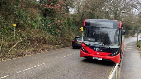 A red bus with a Transport for Cornwall logo on the front drives along a damp Cornish road with trees and hedgerow on one side and a pavement on the other.