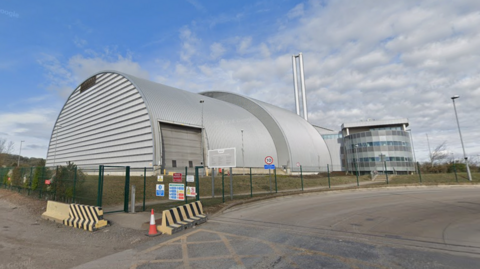A large grey industrial building with an attached three-storey building with windows. The site has a tall, thin double chimney.