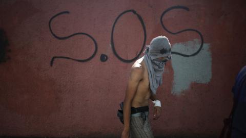A protestor stands, with his head wrapped up, in front of graffiti that spells out SOS