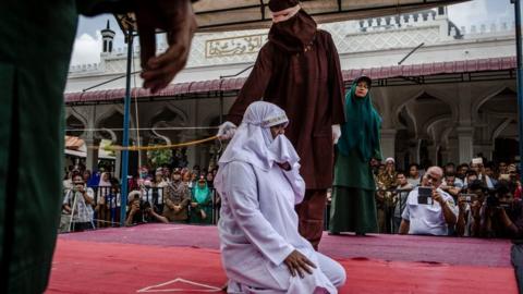 A woman on her knees is flogged in Aceh state by a hooded man