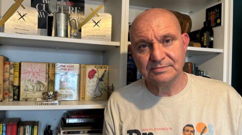 Mike Craven is standing in front of his bookcase and looking at the camera. He is bald and is wearing a 'Ian Fleming's Dr. No' t-shirt. Two gold dagger awards are displayed, along with some of Fleming's books.