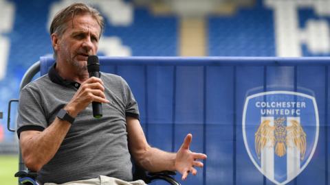 Colchester owner Robbie Cowling at the club's stadium, holding a microphone in his hand and speaking while seated