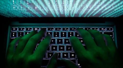 Close up of the hands of a man typing on a computer keyboard. It's very dark, but the keyboard is lit up with data scrolling through the screen