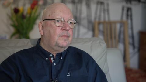 John Kiely sitting on a sofa in his room. He is bald and wears glasses and a long-sleeved navy blue shirt.