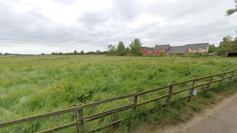 A patch of land with a fence. There are some houses in the background.