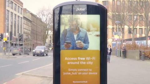 The image shows a smart hub in the foreground which looks like a oversized phone screen. There is a black edge which has the logo 'Pulse' at the top. The screen has a picture of a man looking at his phone in a denim shirt and headphones. Below it says 'Access free wifi around the city. Simply connect to pulse_hub on your device'. Behind the hub is a road with cars and a wide pavement with people. Buildings are visible on both sides.