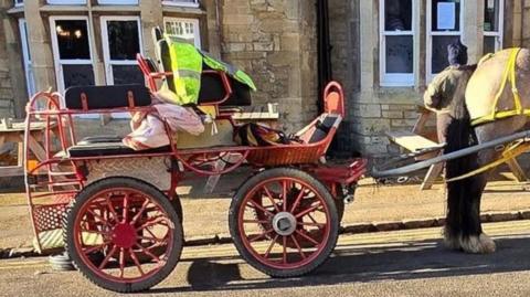 Four-wheeled cart with red and silver paintwork and black seats. One seat is higher than the main body of the vehicle, and has a hi-vis jacket on top of it. It is connected to a black and grey horse with yellow reins and is parked outside a stone-built pub.