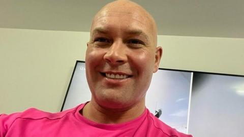 Head and shoulders image of Kirk Marsden, He is bald, smiling and wearing a bright pink tshirt