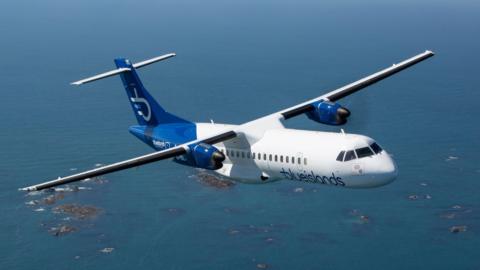 A Blue Islands airplane airborne in the sky with the sea and rocks seen down below.