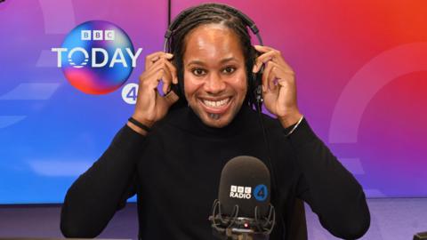 Holding headphones on his head, BBC Radio 4 Today Christmas guest editor Prof Jason Arday smiles at the camera in the studio.