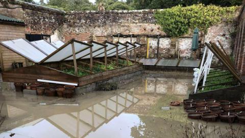A large pool of  brown coloured muddy water surrounds some cold frames. The glass windows are open and green plants can be seen inside.
