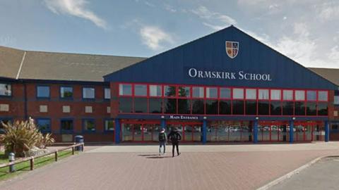 Street view image of Ormskirk School, a two storey red brick building with blue framed windows and a large blue triangular main entrance with a shield crest and 'Ormskirk School' in white. To the front is a red brick paved area with two students walking towards the school