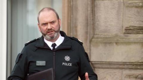 Mark McEwan talking beside a building wall, carrying a black folder. He is wearing a shirt and tie under a black police jacket.