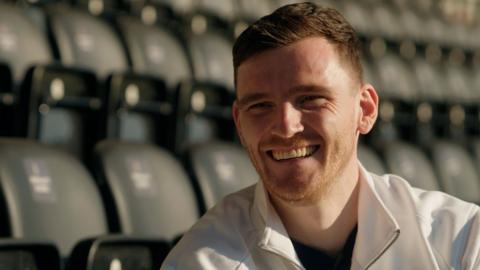 Andy Robertson smiles while sat in football stands - he has short brown hair and wears a white zip jacket