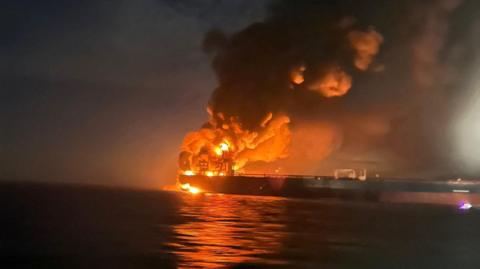 A large tanker with orange flames raging and smoke spiralling, in the night.