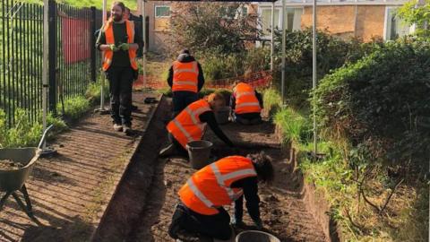 Four people in high vis can be seen kneeling on the ground of an excavation site with buckets next to them. Two other people are walking next to it. A wheelbarrow can be seen in the left corner of the picture, filled with dug up earth.