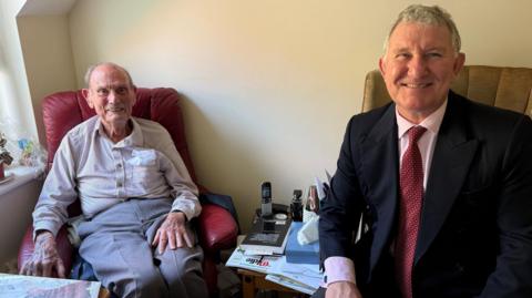 An older gentleman in grey trousers and light coloured shirt sits in a red leather armchair on the left-hand side, smiling at the camera. On the right-hand side a man with grey hair smiles at the camera, wearing a black suit jacket, pink shirt and red tie.