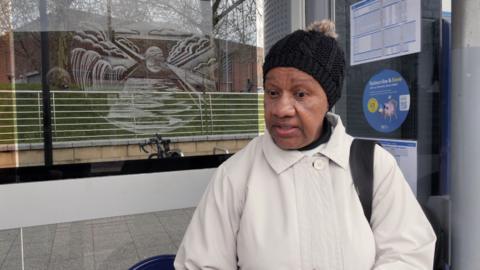 A woman in a white coat and black bobble hat talking at a tram stop
