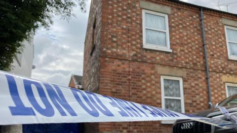 Police tape which says 'police line do not enter' in blue writing, in front of a terraced house with a black Audi car in front.