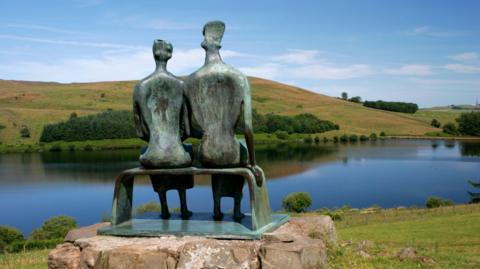 The Henry Moore sculpture King and Queen viewed from behind looking over a reservoir with a tree lined hill in the distance