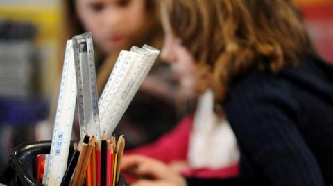 Primary school children at work in a classroom. Their images are blurred in the background. There is container full of pencils and rulers in the foreground.