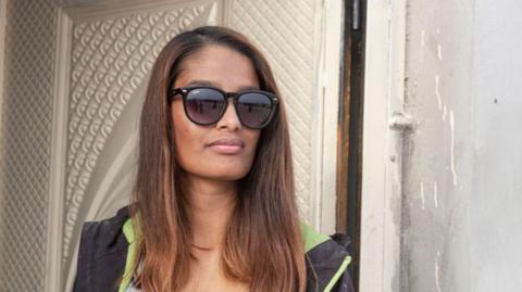 Shamima Begum leaning against a door frame. She has long dyed brown hair and is wearing sunglasses with a light grey top underneath a sweatshirt jacket