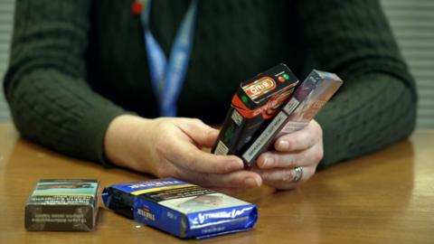 A pair of hands holding two packets of illicit cigarettes. Two more lie on the table.