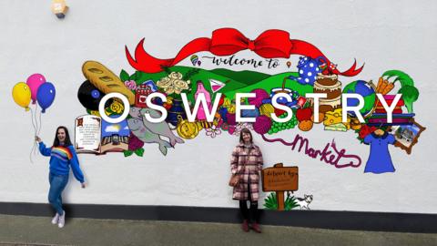 Two women standing in front of a wall with an artist's impression of the colourful mural, featuring the word Oswestry and items that can be bought in the market