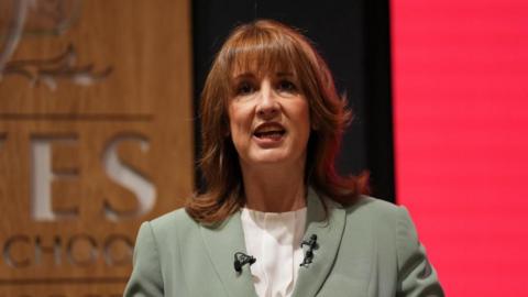 Rachel Reeves wears a sage green blazer while standing in front of a red-lit screen and a wooden sign.