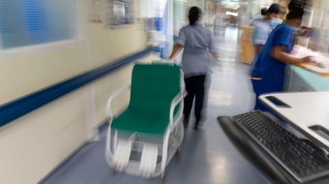 A nurse is moving a hospital chair along a corridor while two other nurses attend to paperwork