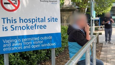 A woman who is wearing a black and light blue jacket and jeans, is sitting smoking outside Hull Royal Infirmary. She is next to a sign which reads 'This hospital site is smoke free.' Her face has been obscured. There is also a man standing a few metres away who is about to light a cigarette. His face has also been blurred. 