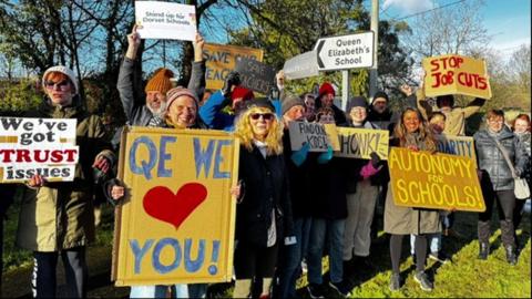 A group of people wearing hats and coats stand on the grass outside QE School holding signs saying "QE we love you" and "Stop job cuts".
