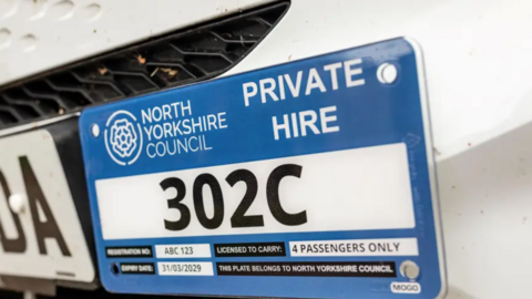 An image of a North Yorkshire Council hackney carriage licencing plate. The plate has a thick blue border with the council's white rose logo on the top left. 
