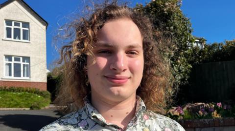 A teenager with curly long hair is standing in front of a house with blue sky in the background. He is wearing a green vintage shirt. 