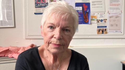 A woman with short grey hair staring down the camera lens with a straight face. wearing a black v-neck t-shirt and a silver necklace, with a notice board with various notices on it in the background.
