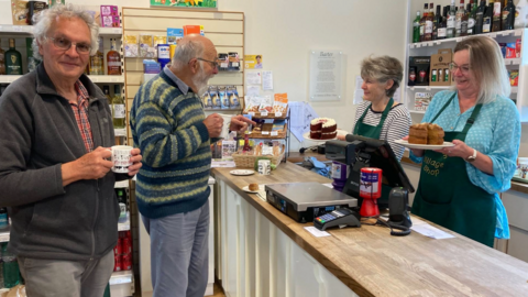 The picture shows the interior of a small shop with two customers holding mugs and two staff members holding plates with cakes. The counter is stocked with items like a scale, cash register, and packaged goods, while shelves behind it display bottles and other products. 
