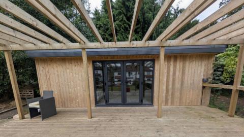 A wooden shed with a deck that has two glass doors and window next to each side of the door. A reflection shows a house on the opposite side and parts of the garden. There is a single grey chair in the corner of the shed.