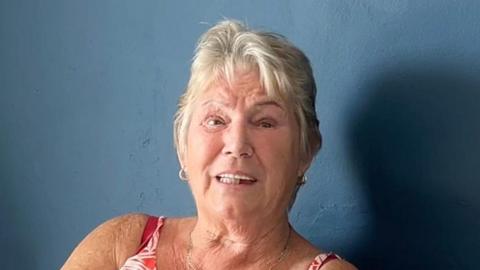 Screengrab of Norma Booth talking while sitting in front of a blue wall. She has short white hair and wears hooped earrings and a red strapped top
