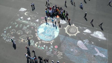 An ariel view of the artwork on Plymouth Hoe. There is Earth at the centre, with 'Happy 100 Birthday Sir David Attenborough' on it. There is a whale to Earth's right and trees and mountains to its left. A choir is gathered at the top the artwork, with people lined up taking photos on the other side.
