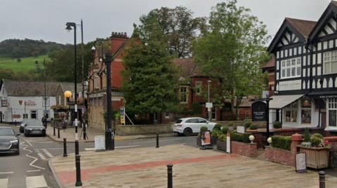 A general street view of Whalley showing green hills in the distance, a pub in front of them, a red brick building to the right partly hidden by trees and a large pavement with bollards in front of a black and white tudor-looking shop