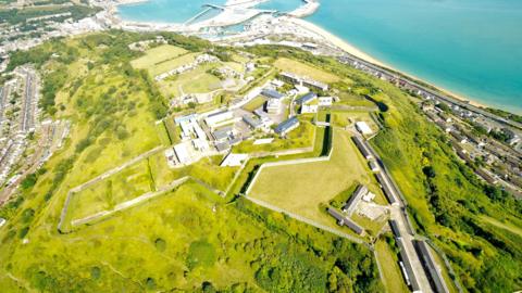 Dover Citadel stands on the cliffs overlooking the port and the English Channel. The site has a mixture of buildings of different ages. The are walls around the site with areas of grass and there is a bridge leading to it.