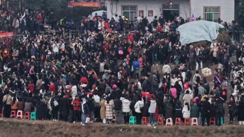 Hundreds of people can be seen standing on the streets outside a white bricked house. The picture is taken from a distance.