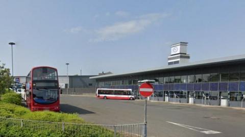 Stanley Bus Station is a long, glass building with a clock tower dominating the roofline in the centre. There are two buses parked in the area outside, and a council vehicle to the right.