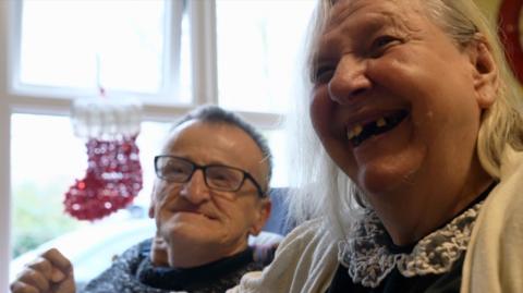 Barry has glasses and short grey hair. He is sitting next to his fiancée Betty who is grinning. There is a red and white sparkly Christmas stocking hanging on the window next to them 