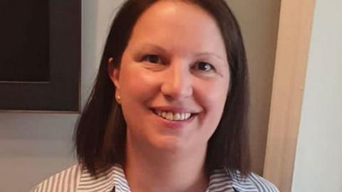 A woman wearing a striped shirt is smiling at the camera. She has brown hair and is standing indoors with what appears to be a television screen attached to a wall behind her.