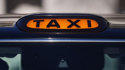 A general view of a taxi sign on top of a black cab. The taxi sign is orange with black font. 