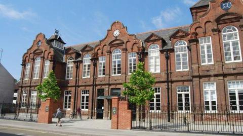 The outside of one of the Norwich University of the Arts buildings in Norwich. There are black railings outside the front of the building and people walking past it.