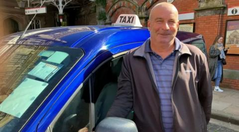 A man with very short grey hair is wearing a blue striped t-shirt and a purple-brown jacket. He is standing next to a blue car that has a sign on the top reading "taxi"