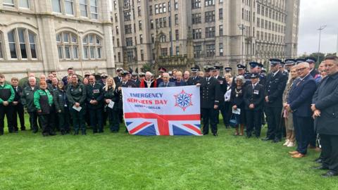 Group of emergency services workers hold Emergency Services Day flag in front of the Liver building