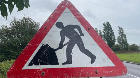 A triangular roadworks street sign showing a black silhouette of a man with a shovel bending over on a white background. The sign has a red edging. Behind it is a glimpse of a road, grass and trees against a grey sky. 